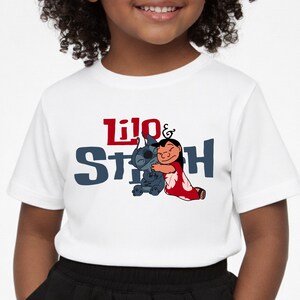 K&ouml;nnte beinhalten: Ein wei&szlig;es kurz&auml;rmeliges T-Shirt mit einem Grafikdruck der sich umarmenden Figuren Lilo und Stitch. Der Schriftzug "Lilo & Stitch" ist in roten und dunkelblauen Buchstaben auf der Brust zentriert &uuml;ber der Zeichentrickillustration gedruckt.