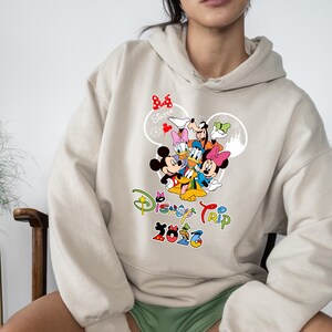 May include: Beige hoodie with a colourful Disney character graphic, including Mickey Mouse, Donald Duck, and Goofy. The text "Disney Trip 2023" is displayed in a vibrant, multi-coloured font, with the word "Sister" above the characters.