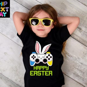 May include: A black t-shirt with a white bunny rabbit wearing a video game controller. The controller is yellow, blue, and black. The text "Happy Easter" is printed in green below the controller.