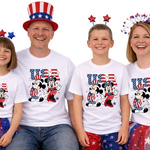 May include: A family wearing matching white t-shirts featuring Mickey and Minnie Mouse in patriotic outfits. The shirts display the word "USA" in red, white, and blue. Some family members are wearing patriotic accessories and holding American flags.