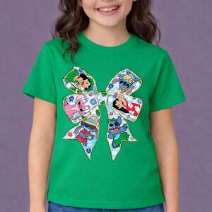 May include: Green t-shirt with a large bow design featuring cartoon characters. The bow is decorated with floral patterns and images of Stitch and other characters. The shirt is a bright green colour.