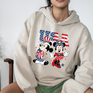 May include: Beige hoodie with a graphic of Mickey and Minnie Mouse in patriotic attire, with the word "USA". Mickey is in a stars and stripes outfit. Minnie wears a red polka dot dress and bow. The design uses red, white, and blue.