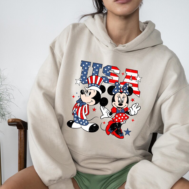 May include: Beige hoodie with a graphic of Mickey and Minnie Mouse in patriotic attire, with the word "USA". Mickey is in a stars and stripes outfit. Minnie wears a red polka dot dress and bow. The design uses red, white, and blue.
