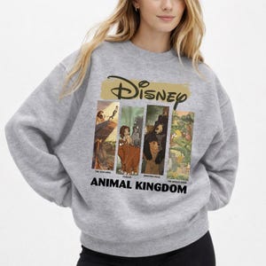 Peut inclure: Un sweat-shirt gris avec un motif Disney. Le design comprend des images du "Roi Lion", de "Tarzan", de "Fr&egrave;re des ours" et du "Livre de la jungle". Le mot "Disney" est au-dessus des images, et "Animal Kingdom" est en dessous.