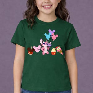 Op de afbeelding: Een donkergroen t-shirt met een stripfiguur die bloemen en ballonnen vasthoudt. De afbeelding bevat een appel met snoep, suikerspin, een cupcake en een ijsje. Het personage is roze met grote ogen.