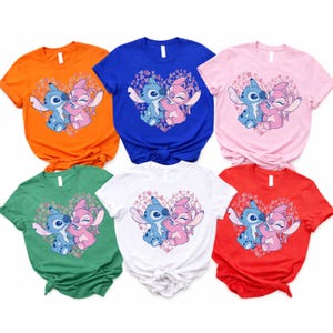 以下が含まれることがあります： オレンジ、青、ピンク、緑、白、赤の6色の半袖Tシャツ。各シャツには、ハート型のデザインの中で抱き合う2人の漫画のキャラクターのグラフィックが描かれています。キャラクターは青とピンクです。