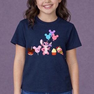 Op de afbeelding: Marineblauw T-shirt met een roze stripfiguur met bloemen en ballonnen. Het shirt heeft ook cartoonafbeeldingen van een appel, suikerspin, een cupcake en een ijsje. Het shirt is voor kinderen.