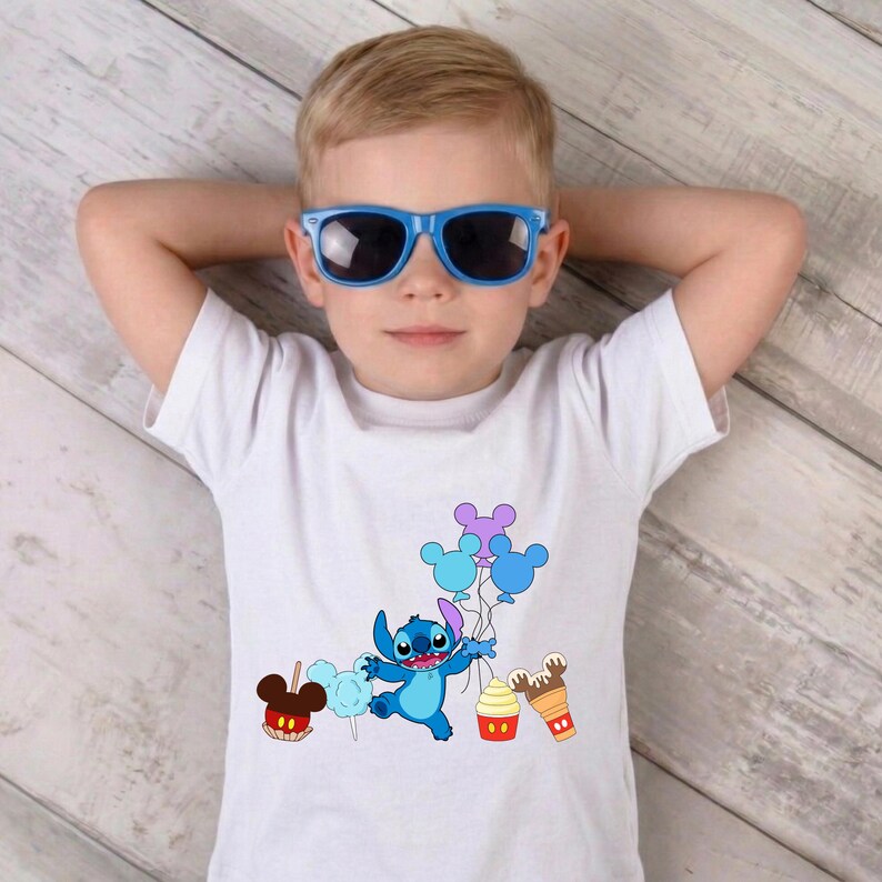 Op de afbeelding: Een wit t-shirt met een blauw stripfiguur dat ballonnen en lekkernijen vasthoudt. Het figuur is omringd door een appel, suikerspin, een cupcake en een ijsje. De jongen draagt een blauwe zonnebril.