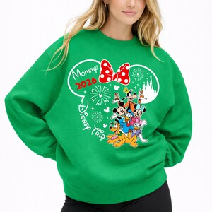 May include: Green sweatshirt featuring a graphic of Disney characters, including Mickey Mouse, Minnie Mouse, Donald Duck, and Goofy. The design includes a red polka-dot bow, fireworks, and the text "Mommy 2026 Disney Trip."