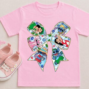 May include: Pink t-shirt with a large bow design featuring Stitch and Lilo characters. The bow is filled with colourful images of the characters. Pink trainers, hair clips, and a hair tie are also in the image.