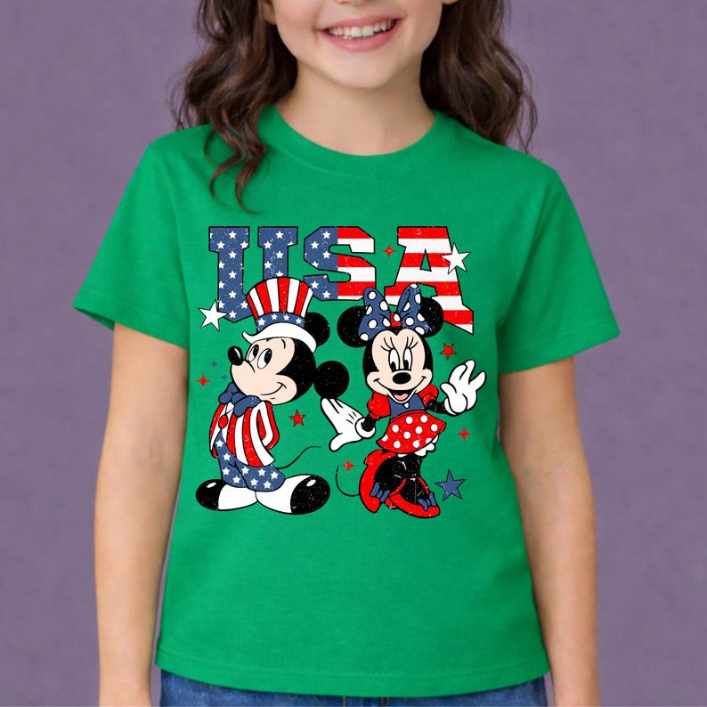 May include: Green t-shirt with a graphic of Mickey and Minnie Mouse in patriotic attire, with the word "USA" in red, white, and blue. The design includes stars and stripes, ideal for celebrating the Fourth of July.