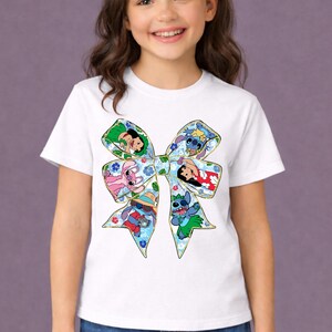 May include: White t-shirt with a large bow design featuring cartoon characters. The bow is decorated with floral patterns and outlines of Stitch, Angel, Lilo, and other characters. The shirt is worn by a young girl.
