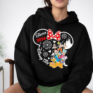 May include: Black hooded sweatshirt featuring a graphic of Disney characters, including Mickey Mouse, Minnie Mouse, and Donald Duck. The design includes a red bow, fireworks, and the text "Sister 2026 Disney Trip."