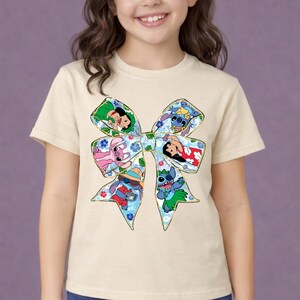May include: A cream-coloured t-shirt featuring a large bow design with various cartoon characters. The bow has a blue floral pattern and contains images of Stitch and other characters. The shirt is worn by a young person.