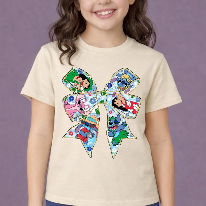 May include: A cream-coloured t-shirt featuring a large bow design with various cartoon characters. The bow has a blue floral pattern and contains images of Stitch and other characters. The shirt is worn by a young person.