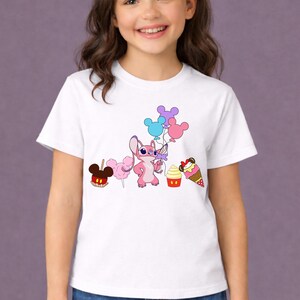 Op de afbeelding: Wit T-shirt met een roze cartoonfiguur met ballonnen en cartoon traktaties. De traktaties zijn onder meer een Mickey Mouse-vormige appel, suikerspin, een cupcake en een ijsje. Het T-shirt wordt door een jong meisje gedragen.