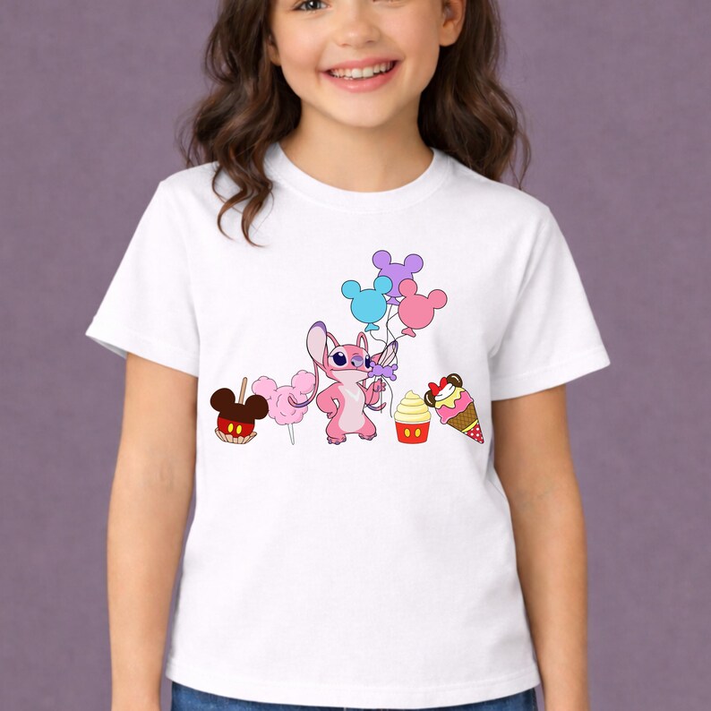 Op de afbeelding: Wit T-shirt met een roze cartoonfiguur met ballonnen en cartoon traktaties. De traktaties zijn onder meer een Mickey Mouse-vormige appel, suikerspin, een cupcake en een ijsje. Het T-shirt wordt door een jong meisje gedragen.
