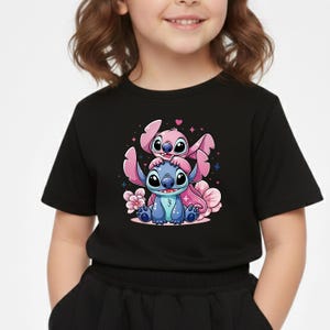 Pode incluir: T-shirt preto com um gr&aacute;fico de desenho animado de duas personagens, uma azul e outra rosa, com flores. O desenho est&aacute; centrado na frente da t-shirt.
