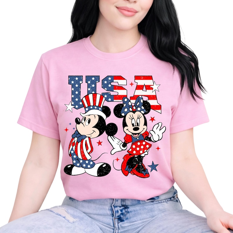 May include: Pale pink t-shirt with a graphic of Mickey and Minnie Mouse in patriotic attire, and the word "USA" in a star-spangled design. The design uses red, white, and blue colours, ideal for celebrating Independence Day.