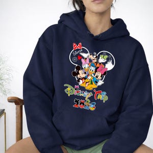 May include: Navy blue hooded sweatshirt with a vibrant graphic of Disney characters, such as Mickey and Minnie Mouse, Donald and Daisy Duck, and Goofy. The text "Disney Trip 2023" is printed below the characters. A red bow is above the word "Sister".