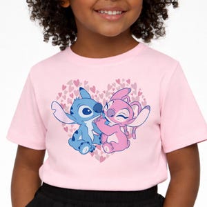 以下が含まれることがあります： 薄いピンク色のTシャツ。小さなハートでできたハートの中で抱き合う2人のキャラクターの漫画のグラフィックが特徴です。一方のキャラクターは青、もう一方はピンクです。このTシャツは半袖です。