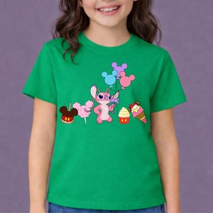 Op de afbeelding: Een groen t-shirt met een cartoonillustratie van een roze personage dat ballonnen vasthoudt, samen met cartoonlekkernijen. De lekkernijen omvatten een Mickey Mouse-vormige appel, suikerspin, een cupcake en een ijsje.
