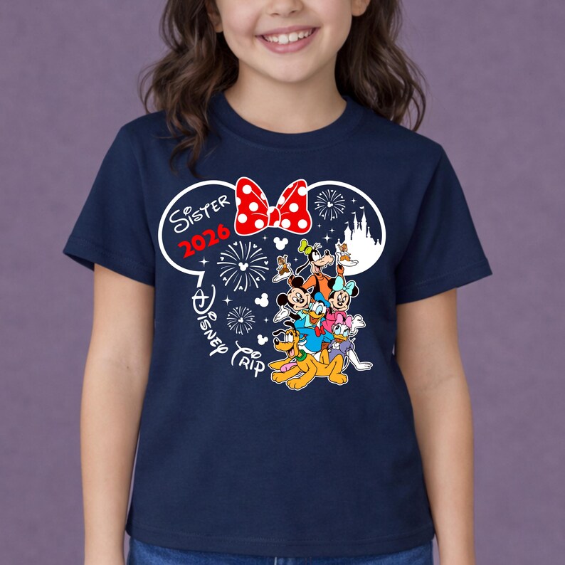 May include: Navy blue t-shirt featuring a graphic of Disney characters, including Mickey Mouse, Minnie Mouse, and Goofy. The design includes a red bow, fireworks, and the text "Sister 2026 Disney Trip". The shirt is worn by a child.