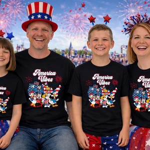 May include: A family of four wearing matching black t-shirts with the text "America Vibes" and a cartoon graphic. The background includes fireworks, American flags, and festive decorations. The family is smiling and holding American flags.