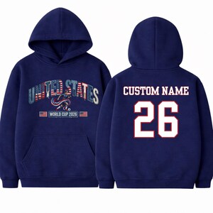 Peut inclure: Sweat-shirt &agrave; capuche bleu marine avec l'inscription "UNITED STATES", un graphisme de joueur de football et le texte "WORLD CUP 2026". Le dos affiche "CUSTOM NAME" et le num&eacute;ro "26" en blanc avec une bordure rouge.