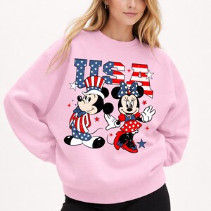May include: A pale pink sweatshirt with Mickey and Minnie Mouse in patriotic outfits. Mickey is in a hat and waistcoat with a star-spangled design, and Minnie wears a polka-dot skirt and bow. The word "USA" is printed above them in red, white, and blue.