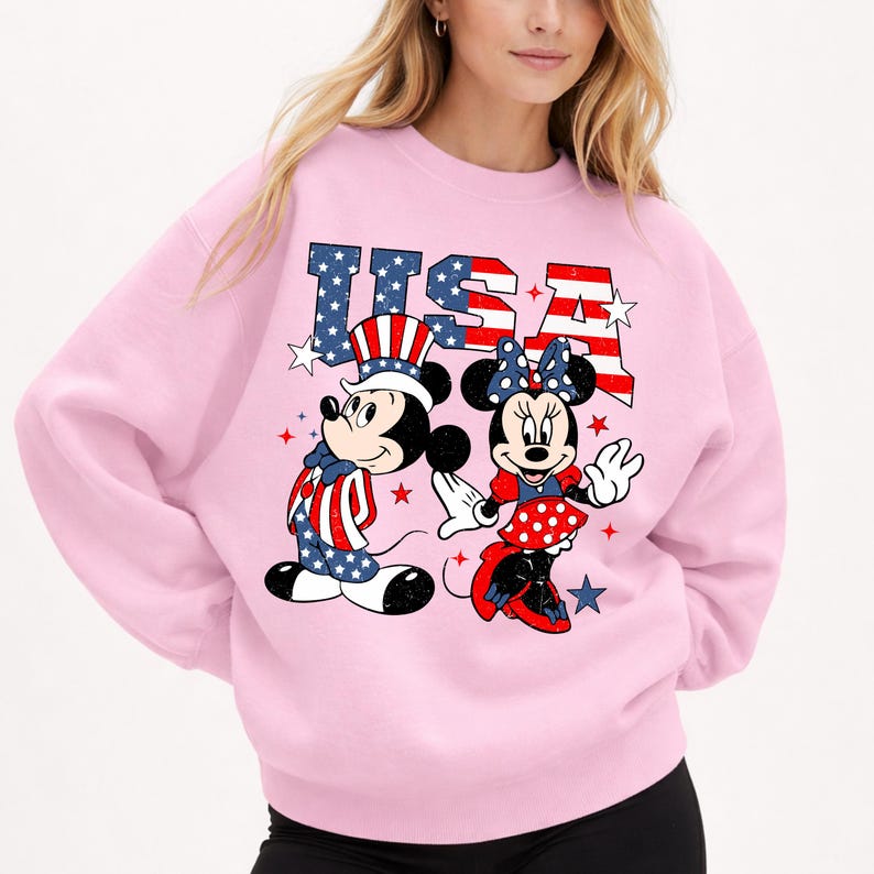 May include: A pale pink sweatshirt with Mickey and Minnie Mouse in patriotic outfits. Mickey is in a hat and waistcoat with a star-spangled design, and Minnie wears a polka-dot skirt and bow. The word "USA" is printed above them in red, white, and blue.