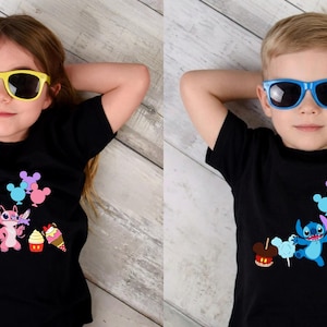 Op de afbeelding: Twee kinderen dragen zwarte t-shirts met een cartoonkarakterontwerp en zonnebrillen. De shirts hebben een kleurrijke afbeelding van een personage, ballonnen en lekkernijen. Het kind links draagt een gele zonnebril, het kind rechts een blauwe zonnebril.
