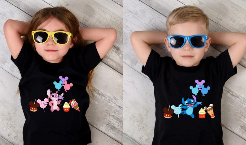 Op de afbeelding: Twee kinderen dragen zwarte t-shirts met een cartoonkarakterontwerp en zonnebrillen. De shirts hebben een kleurrijke afbeelding van een personage, ballonnen en lekkernijen. Het kind links draagt een gele zonnebril, het kind rechts een blauwe zonnebril.
