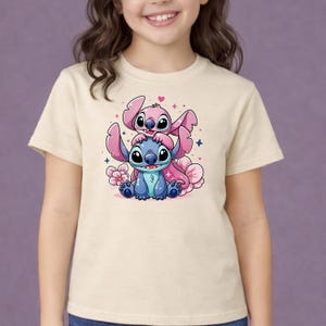 Pode incluir: Uma camiseta bege com um desenho animado colorido de dois personagens, um azul e um rosa, com olhos e orelhas grandes. O design inclui flores e cora&ccedil;&otilde;es rosa, criando uma est&eacute;tica l&uacute;dica e caprichosa.