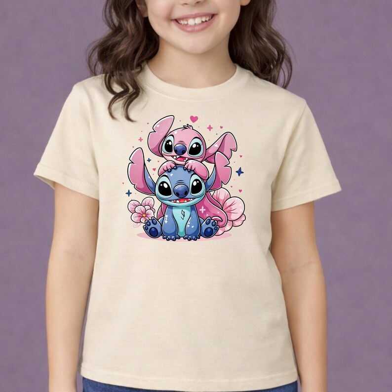 Pode incluir: Uma camiseta bege com um desenho animado colorido de dois personagens, um azul e um rosa, com olhos e orelhas grandes. O design inclui flores e cora&ccedil;&otilde;es rosa, criando uma est&eacute;tica l&uacute;dica e caprichosa.