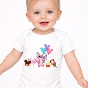 Op de afbeelding: Witte babybody met een roze stripfiguur met ballonnen, samen met een Mickey Mouse-vormige appel, suikerspin, een cupcake en een ijsje. De baby lacht en heeft blonde staartjes.