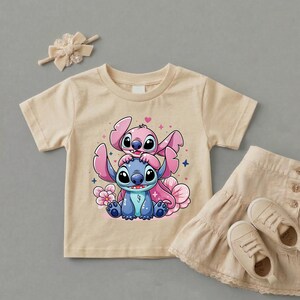 Pode incluir: Uma camiseta bege para crian&ccedil;as pequenas com uma ilustra&ccedil;&atilde;o de desenho animado de Stitch, um alien&iacute;gena azul, e Angel, um alien&iacute;gena rosa, com flores e cora&ccedil;&otilde;es rosa. Uma saia, sapatos e um la&ccedil;o combinando tamb&eacute;m est&atilde;o na imagem.