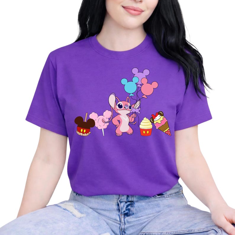 Op de afbeelding: Een paars T-shirt met een stripfiguur die ballonnen vasthoudt en omringd is door snoep. De afbeelding bevat een appel met snoep, suikerspin, een cupcake en een ijsje. Het ontwerp is in cartoonstijl.