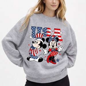 May include: A grey sweatshirt with Mickey and Minnie Mouse in patriotic outfits. Mickey is in red, white, and blue. Minnie wears a red polka-dot dress and bow. The word "USA" is printed above them in red and blue, with star details.