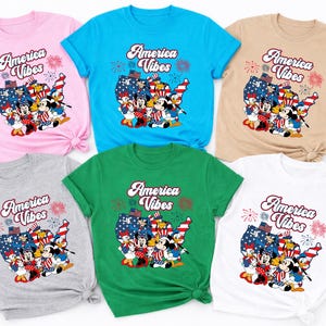 May include: A collection of six t-shirts in various colors: pink, blue, tan, gray, green, and white. Each shirt features the text "America Vibes" above a graphic of cartoon characters in patriotic attire, set against a map of the United States.