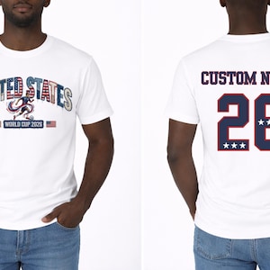 Peut inclure: T-shirt blanc avec "UNITED STATES" dans un design patriotique, avec un graphisme de joueur de football et "WORLD CUP 2026". Le dos affiche "CUSTOM NAME" au-dessus du num&eacute;ro 26 avec des &eacute;toiles.