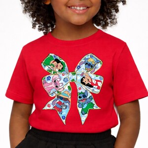 May include: A red t-shirt with a large bow design featuring cartoon characters. The bow has a blue floral pattern and contains images of various characters. The short-sleeved shirt is likely made of cotton.