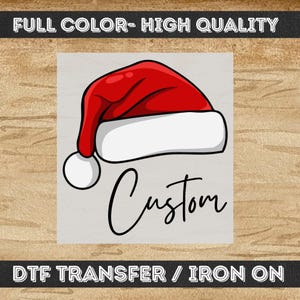 May include: A red and white Santa hat graphic with a white pom-pom, positioned above the word Custom in black cursive script. The image includes the text FULL COLOR- HIGH QUALITY at the top and DTF TRANSFER / IRON ON at the bottom.