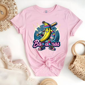 K&ouml;nnte beinhalten: Pinkes T-Shirt mit einer Cartoon-Banane, die einen Baseballschl&auml;ger h&auml;lt und eine Baseballkappe tr&auml;gt. Die Banane tr&auml;gt eine rosa-blaue Uniform. Der Text "Bananas" ist in einer Retro-Schriftart geschrieben.