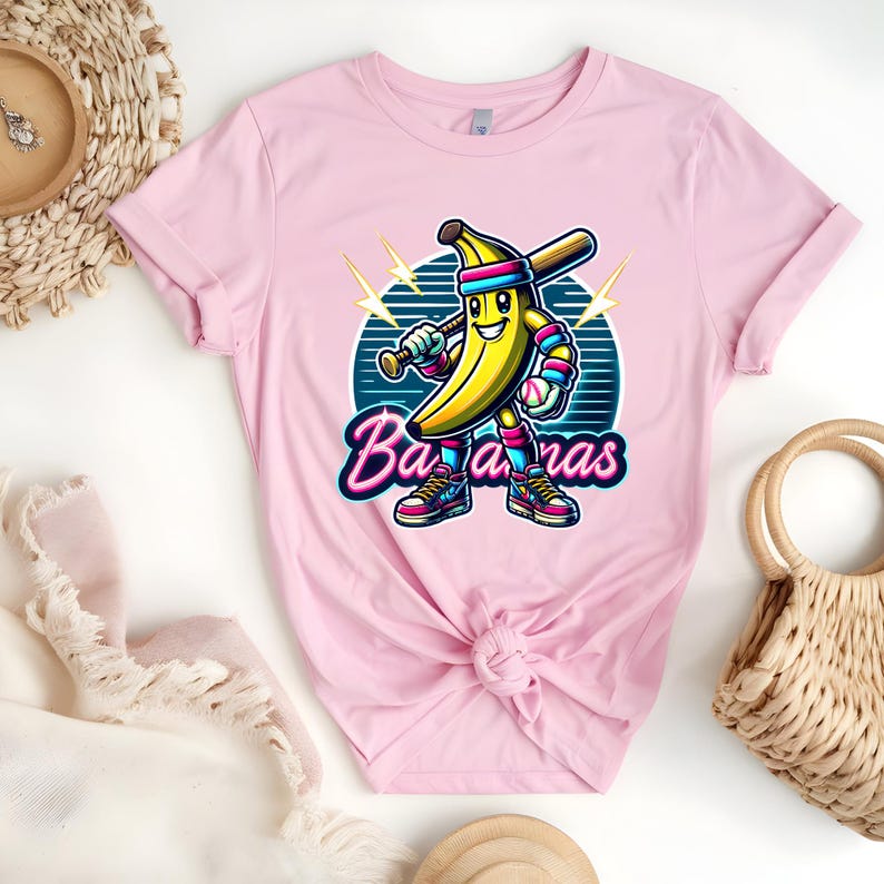 K&ouml;nnte beinhalten: Pinkes T-Shirt mit einer Cartoon-Banane, die einen Baseballschl&auml;ger h&auml;lt und eine Baseballkappe tr&auml;gt. Die Banane tr&auml;gt eine rosa-blaue Uniform. Der Text "Bananas" ist in einer Retro-Schriftart geschrieben.