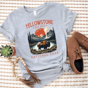 May include: A light gray t-shirt featuring a graphic of Yellowstone National Park. The design includes a bison, mountains, a lake, and the text "YELLOWSTONE EST. 1872 NATIONAL PARK" in red. Brown shoes are visible on the right.