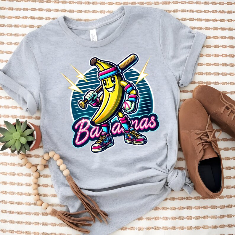 K&ouml;nnte beinhalten: Ein graues T-Shirt mit einer Cartoon-Banane, die ein Baseball-Uniform tr&auml;gt und einen Schl&auml;ger h&auml;lt. Die Banane tr&auml;gt ein rosa Stirnband und hat einen gelben und blauen Baseball. Der Text "Bananas" ist in pinken Neonbuchstaben.