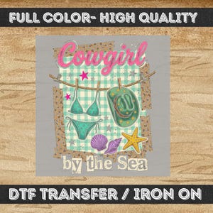 Peut inclure: Conception graphique avec un bikini turquoise, un chapeau de cow-boy et des coquillages suspendus à une corde à linge. Les mots "Cowgirl" et "by the Sea" sont affichés en rose et blanc sur un fond gris.