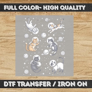 May include: A DTF transfer iron-on featuring a space-themed pattern with six cats wearing glass astronaut helmets. The cats are depicted in various colors including orange tabby, black, and calico, floating among white line-art planets and stars.