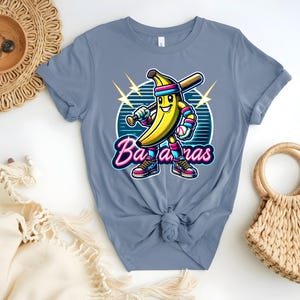 K&ouml;nnte beinhalten: Ein hellblaues T-Shirt mit einer Cartoon-Banane, die einen Baseballschl&auml;ger h&auml;lt und eine Baseballkappe tr&auml;gt. Die Banane tr&auml;gt Turnschuhe und hat ein L&auml;cheln im Gesicht. Der Text "Bananas" ist in Neon-Pink und Blau geschrieben.